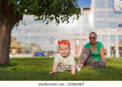 Baby Crawling On The Grass On A Background Of Sitting His Mom. The Kid Runs Away From His Mother. Mother Playing With Her Child.