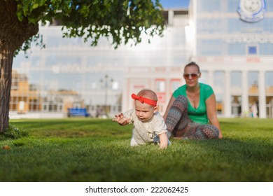 Baby Crawling On The Grass On A Background Of Sitting His Mom. The Kid Runs Away From His Mother. Mother Playing With Her Child.