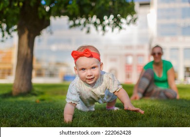 Baby Crawling On The Grass On A Background Of Sitting His Mom. The Kid Runs Away From His Mother. Mother Playing With Her Child.