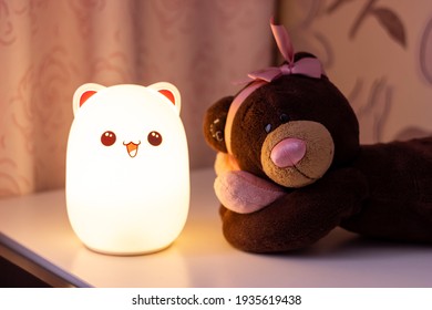 The baby cute bear-shaped night lamp with eyes and ears near a toy teddy bear with a bow on its head and a heart in its hands on the bedside table in the pink-style room glows in the dark