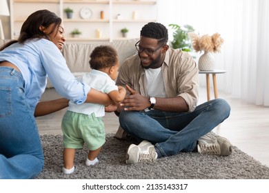 Baby Development Concept. Little Black Infant Boy Making First Steps At Home, Happy Mum Helping Toddler Kid Walking To Dad, Young Parents Spending Time With Child In Living Room Sitting On Floor