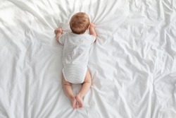 Baby Development Concept. Top View Of Infant Child Crawling On Bed, Newborn Boy Or Girl In Diaper And Striped Bodysuit Relaxing In White Bedroom, Small Little Kid Lying On His Tummy, Copy Space