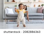 Baby Development. Cute Little Black Infant Boy Making First Steps At Home, Adorable African American Toddler Child Walking In Living Room, His Happy Mom Smiling On Background, Copy Space