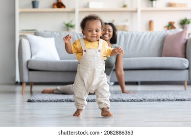 Baby Development. Cute Little Black Infant Boy Making First Steps At Home, Adorable African American Toddler Child Walking In Living Room, His Happy Mom Smiling On Background, Copy Space