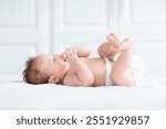 Baby in diaper in white sunny bedroom. Newborn child relaxing in bed. Nursery for young children. Textile and bedding for kids. Family morning at home. New born kid in nappy.