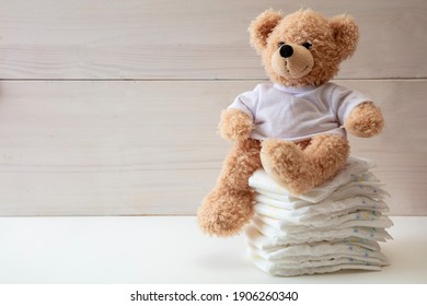 Baby diapers stack cute teddy bear sitting on top, white color floor, wooden wall background. Newborn nappies concept