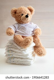 Baby diapers stack, cute teddy bear sitting on top, white color floor, wooden wall background. Newborn nappies concept