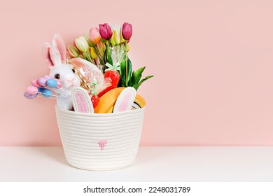 Baby Easter Basket gifts. White  Easter Basket with Easter Bunny, eggs, flowers, candy and toys on pink background. Holidays decorations.  