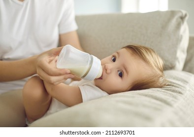 Baby enjoying nutritious meal. Mother feeding baby on sofa. Nanny formula feeding little child as alternative to breastfeeding. Baby boy or girl lying on couch and drinking milk from plastic bottle - Powered by Shutterstock