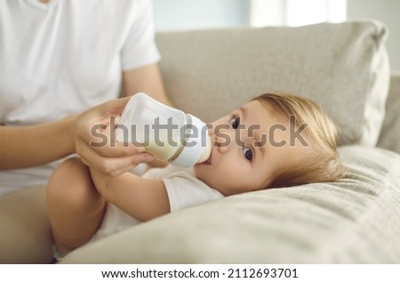 Baby enjoying nutritious meal. Mother feeding baby on sofa. Nanny formula feeding little child as alternative to breastfeeding. Baby boy or girl lying on couch and drinking milk from plastic bottle