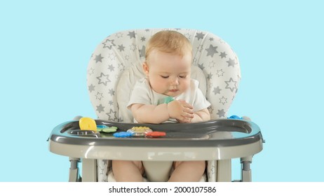 Baby In The Feeding Chair Front View. A Six-month-old Baby Is Sitting On A High Chair Playing With Toys And Pulling Them Into His Mouth