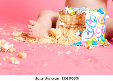 Baby feet in a messy cake that has been smashed for first birthday celebration on a solid pink background. 