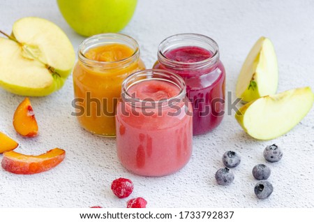 Baby food. Homemade fruit puree. Variety of apple puree or applesauce with frozen peach, raspberries and blueberries in three glass jars on a light background. Healthy food. Horizontal.