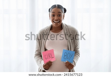 Baby gender reveal concept. Pregnancy celebration with friends. Excited young pregnant black woman holding pink and blue girl and boy cards, standing by window at home. Maternity event.