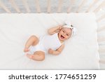 baby girl 6 months old lies in a crib in the nursery with white clothes on her back and laughs, looks at the camera, baby