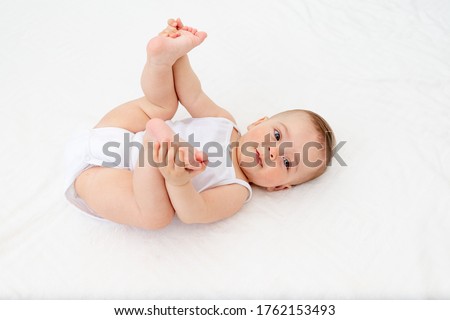 baby girl 6 months old lying in a crib in the children's room on her back and holding her legs, looking at the camera, baby's morning, baby products concept