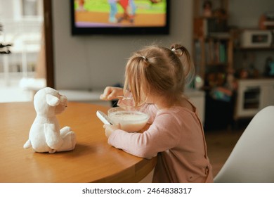 Baby girl eats breakfast and watches cartoons on TV. Children and technology concept. Alpha Generation and modern technology with electronic device. Part of the series. - Powered by Shutterstock