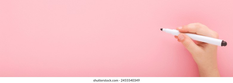 Baby girl hand holding black white color pen and writing on light pink table background. Pastel color. Closeup. Wide banner.  Empty place for text. Top view. - Powered by Shutterstock