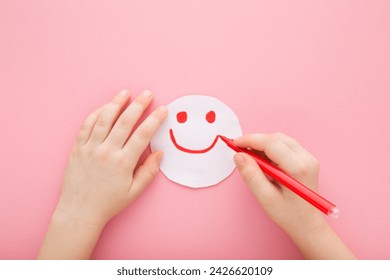 Baby girl hand holding red marker and drawing smiley. Happy smiling face expression on white paper. Light pink table background. Pastel color. Closeup. Point of view shot. - Powered by Shutterstock