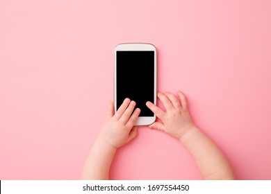 Baby Girl Hands Playing With Smartphone On Light Pink Table Background. Pastel Color. Closeup. Empty Place For Text On Black Screen. Point Of View Shot.