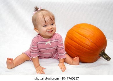 A Baby Girl With Her Hair Tied In A Ribbon On Top Of Her Head Sits Straddling On A White Blanket Next To A Pumpkin.  Her Tongue Is Slightly Out Like She Is Blowing Rasberries.  There Is A Path.