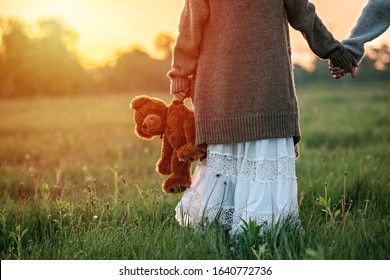 Baby girl holds a teddy bear in hand at dawn in the grass.