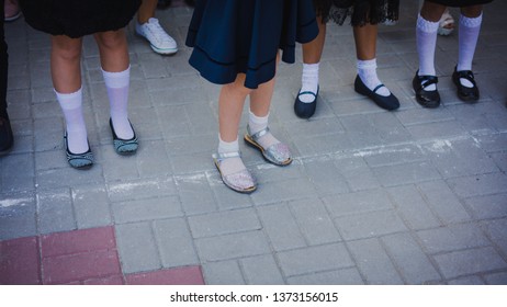 Baby Girl Legs In White Golf And Beautiful Shoes Are In The Lineup On The School Line On September 1. September 1 - Line Schoolyard. First Graders.