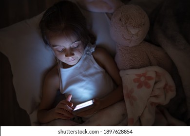 Ein kleines Mädchen liegt mit einem weichen Spielzeug im Bett und benutzt ein Smartphone. Das Konzept der Kinderabhängigkeit von Cartoons und Spielen.