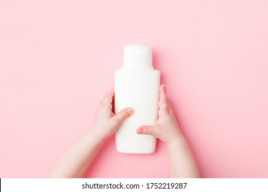 Baby Girl Little Hands Holding White Cream Bottle On Light Pink Table Background. Pastel Color. Care About Clean And Soft Body Skin. Daily Children Beauty Product. Closeup. Point Of View Shot.