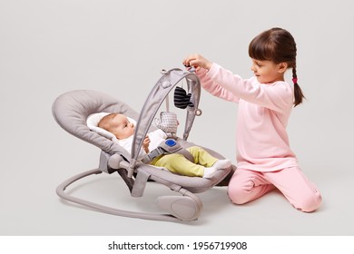 Baby girl lying down in cradle with her sister, little girl with pigtails sitting on floor near her newborn sis, posing isolated over white background.