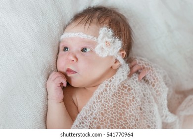 Baby Girl Lying Under A Light Knit Blanket With A Flower On His Head. Baby Girl With Open Eyes.