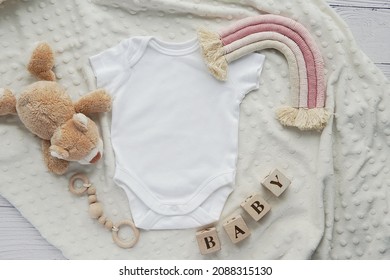 Baby girl onesie mockup, boho rainbow decoration, toys, pregnancy announcement, baby waiting flat lay composition on soft blanket.