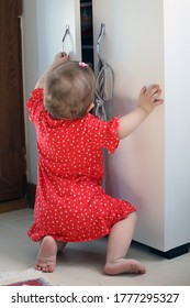 Baby Girl In Red Dress Trying To Open Kitchen Cabinet