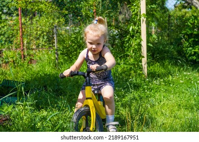 Baby Girl Rides A Running Bike In The Summer On The Lawn At The Dacha.