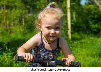 Baby Girl Rides A Running Bike In The Summer On The Lawn At The Dacha.