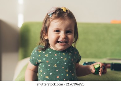 Baby Girl Showing Her White Teeth While Posing Looking At The Camera And Holding A Cucumber. Small House. Happy Family. Closeup Portrait. Kid Life. Beauty