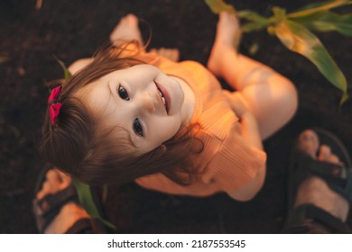 Baby Girl Sitting In Green Cornfield On Sunny Summer Day, Looking Up At Camera. Family Exploring Nature Together. Country Environment.