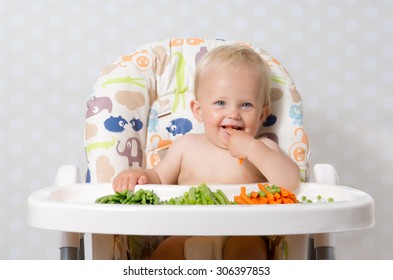 Baby Girl Sitting In A Highchair Eating Raw, Seasonal Vegetables: Carrots, Beans, Peas, Celery