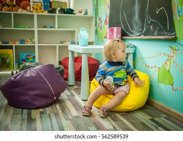 Baby Girl Is Sitting On The Floor. Modern Bright Child Room. Cushioned Furniture, Chair Bag. Happy Child Playing At Home Or Daycare. Educational Toys For Preschool And Kindergarten Child.