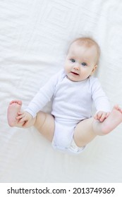 Baby Girl Six Months Old Plays With Her Feet Lying On Her Back On A White Bed In The Bedroom Of The House