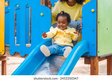 Baby girl sliding in a blue slide in a park with her mother in her back watching and controlling - Powered by Shutterstock