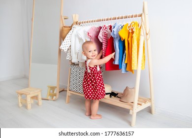 Baby girl stands near a wardrobe and chooses a dress and smiles. Dressing closet with clothes arranged on hangers. Wardrobe of newborn, kids, toddlers, babies full of all clothes. montessori wardrobe.