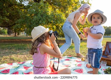 Baby Girl Takes Photos Of Baby Brother With Analog Camera, While Mom Talks On Phone During Picnic.