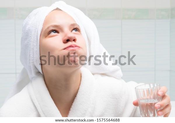 baby girl, in a white robe and towel on her\
head, in the bathroom, holding a glass of water and rinsing her\
mouth, after cleaning