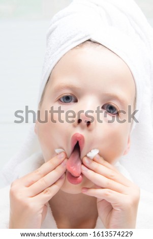 baby girl, with a white towel on her head, fooling around, cleaning her lips with a round cotton swab, after lipstick, close-up, against the wall in the bathroom