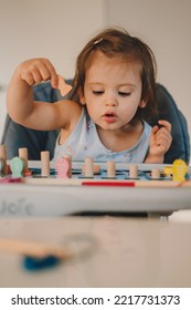 The Baby Girl Wondering Where Is The Place Of The Figurine She Is Holding In Her Hand, On The Wooden Board She Has On The Table. Educational Games. A Light