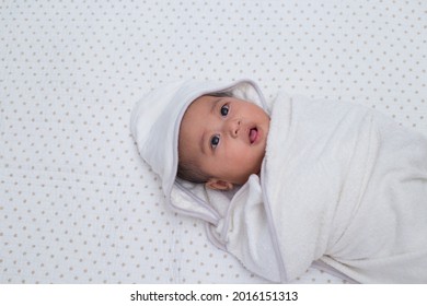 Baby Girl Wrapped In White Towel After Bath, Lying On Bed. Southeast Asian 4 Months Old Daughter On White Background Wrapped, Looking Upward. 