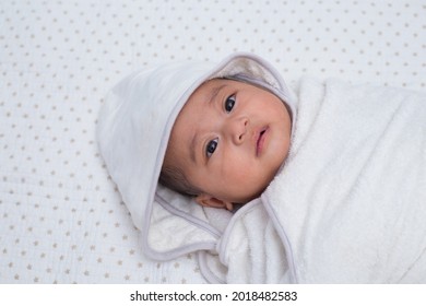 Baby Girl Wrapped In White Towel After Bath, Lying On Bed. Southeast Asian 4 Months Old Daughter On White Background Wrapped, Looking Upward. 