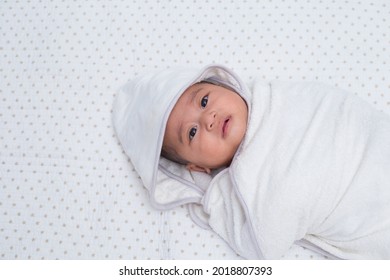 Baby Girl Wrapped In White Towel After Bath, Lying On Bed. Southeast Asian 4 Months Old Daughter On White Background Wrapped, Looking Upward. 