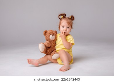 a baby girl in a yellow bodysuit sits and plays with a teddy bear on a white background